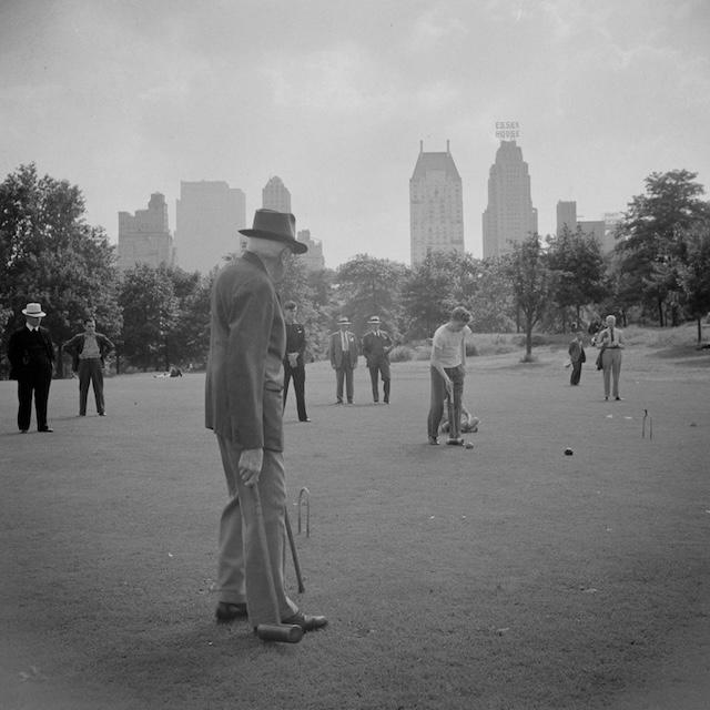 Men playing a game of croquet on the green, with New York City's tall buildings in the background. The game represents a time when leisure activities were more slow-paced and community-oriented