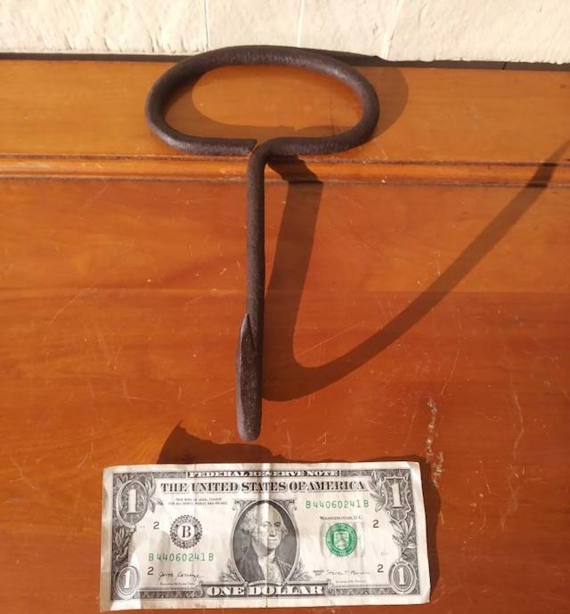 Comparing size to a dollar bill, this photo highlights how compact the antique hay hook is, yet how powerful it was in the hands of those who worked with hay daily