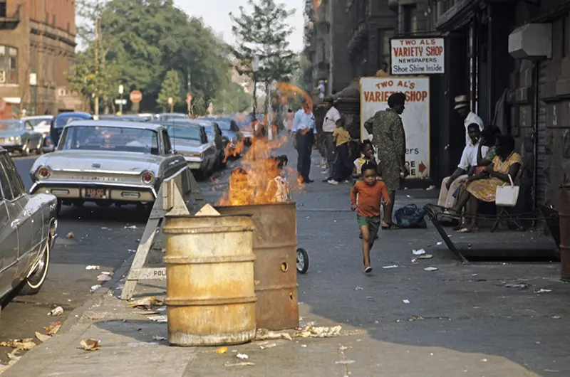 A child walks past a burning trash can in Harlem—a stark symbol of the city’s struggles.