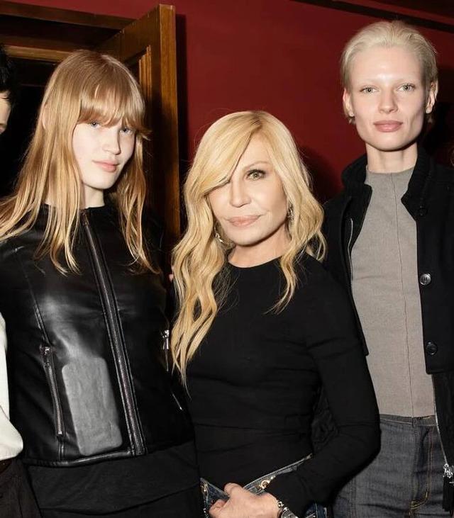 Donatella Versace surrounded by models at an event, sporting her signature style with a chic black top.