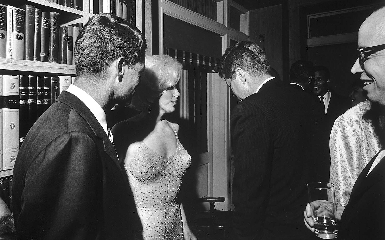 President Kennedy, with his head slightly tilted, listens intently to Marilyn, while his brother, Robert Kennedy, stands nearby, watching the captivating moment.