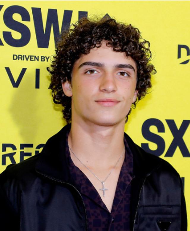 He arrived in a patterned shirt layered under a satin bomber jacket, paired with camel-toned pants that struck the perfect balance between polished and youthful