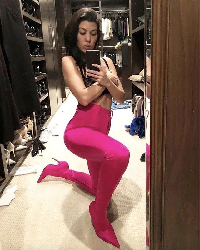 Kourtney Kardashian snapping a mirror selfie in bright pink boots, surrounded by scattered items in her walk-in closet.