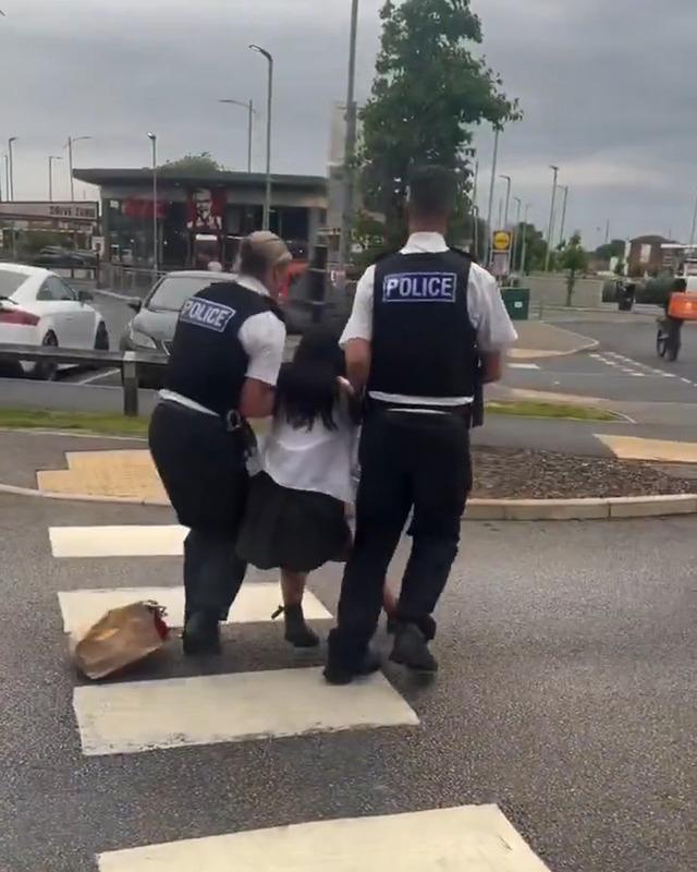 The girl is held between two officers as she is led across the street, leaving her McDonald’s paper bag behind.