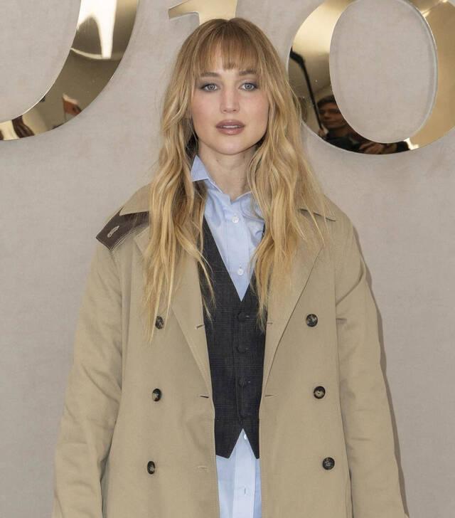 Jennifer Lawrence confidently wearing a classic beige trench coat, drawing attention at Paris Fashion Week amidst the debate over her cosmetic choices.