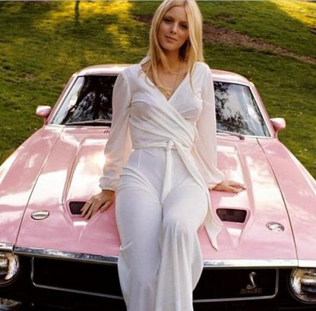 Liv Lindeland exudes classic beauty as she poses atop her pink car, embodying the perfect balance of elegance and adventure.