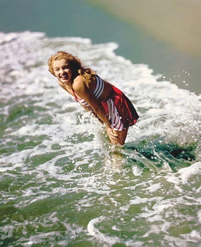 A cheerful moment of Norma Jeane playing at the shoreline, laughing as waves crash around her—one of the most iconic images from her early pinup-style sessions.