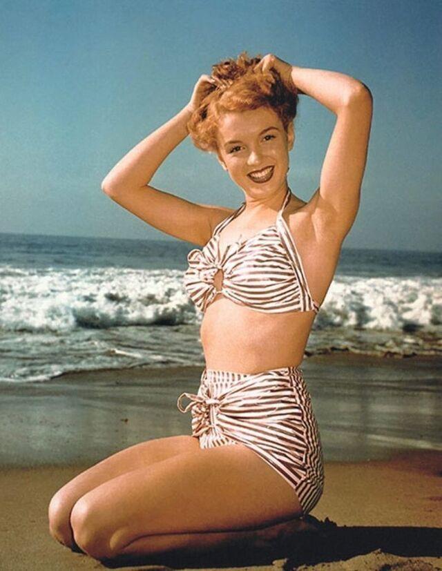 A vibrant beach portrait of teenage Norma Jeane posing playfully in a striped two-piece swimsuit, showcasing her early confidence in front of the camera.