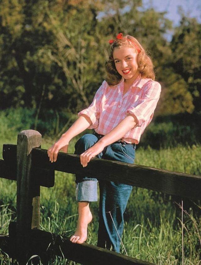 Norma Jeane dressed in a gingham blouse and denim jeans, posing by a wooden fence in a countryside setting—a reminder of her humble, wholesome beginnings.